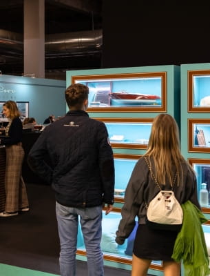 A man and a woman in front of a window displaying luxury maritime products in a chic space with people in the background