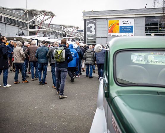Foule entrant dans un salon automobile avec une vieille voiture verte au premier plan.